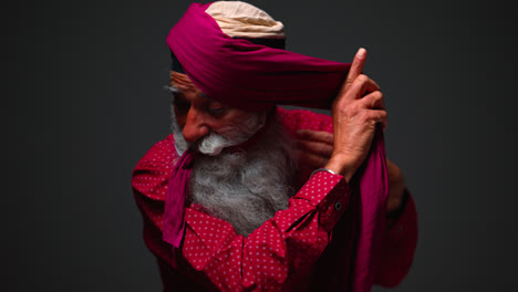 Fotografía-De-Estudio-Con-Iluminación-Tenue-De-Un-Hombre-Sikh-Mayor-Con-Barba-Atando-Una-Tela-Para-Hacer-Un-Turbante-Contra-Un-Fondo-Oscuro,-Filmada-En-Tiempo-Real